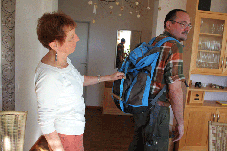 Margit und Achim zuhause, Margit demonstriert, wie sie sich bei Wanderungen am Rucksack festhält