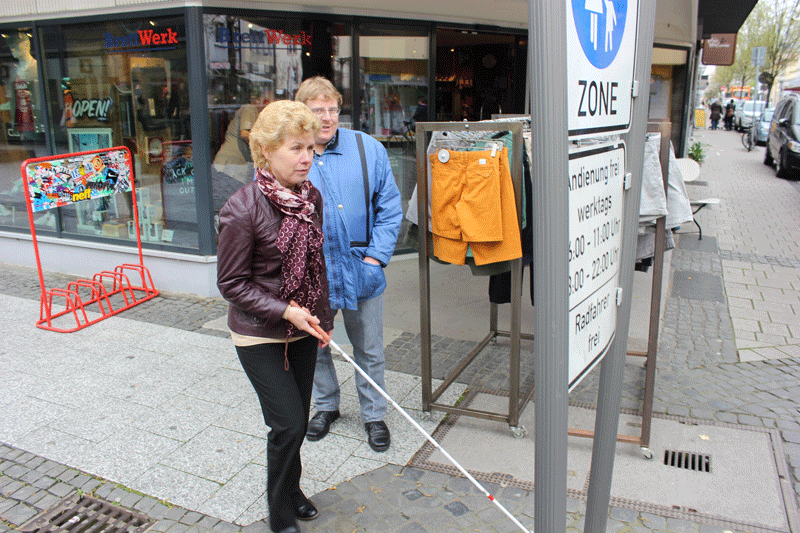Silvia mit ihrem Stock zwischen Kleiderat&auml;ndern und einem Schild, das aud die Fu&szlig;g&auml;ngerzone hinweist