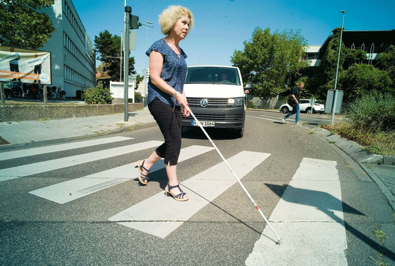 Silvia Sch&auml;fer &uuml;berquert mit demBlindenstock einen Zebrastreifen, dahinter ein Auto