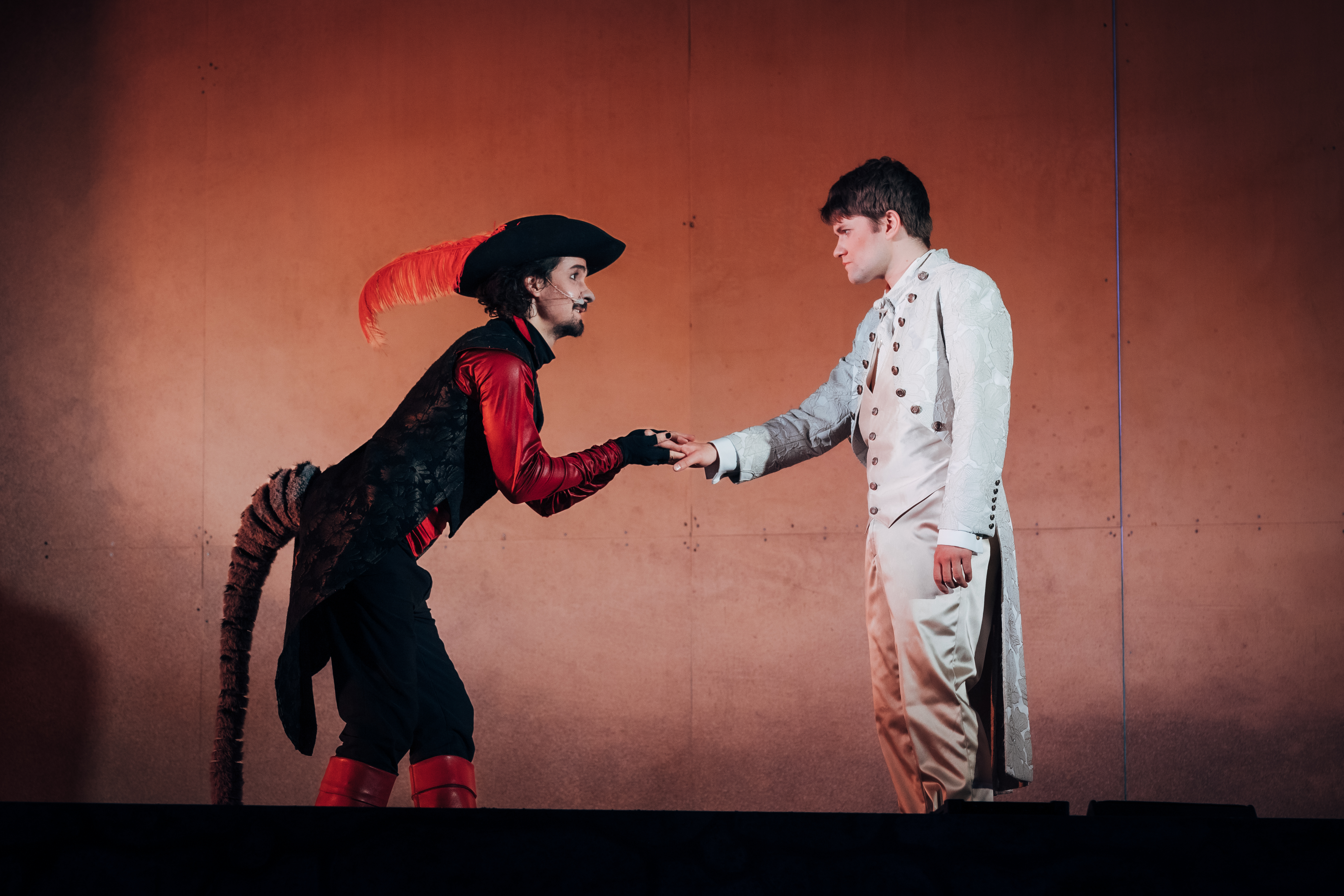 Das Bild zeigt zwei Personen auf einer B�hne. Die linke Person tr�gt ein Theaterkost�m, das einer Katze �hnelt, mit einem schwarzen Hut mit einer gro�en roten Feder, einem roten Hemd, einer schwarzen Weste, einer schwarzen Hose, roten Stiefeln und einem langen Schwanz. Diese Person verbeugt sich leicht. Der Mann auf der rechten Seite tr�gt einen wei�en, gekn�pften Mantel mit passender Hose und schwarzen Schuhen. Er steht aufrecht. Beide Personen geben sich die Hand.