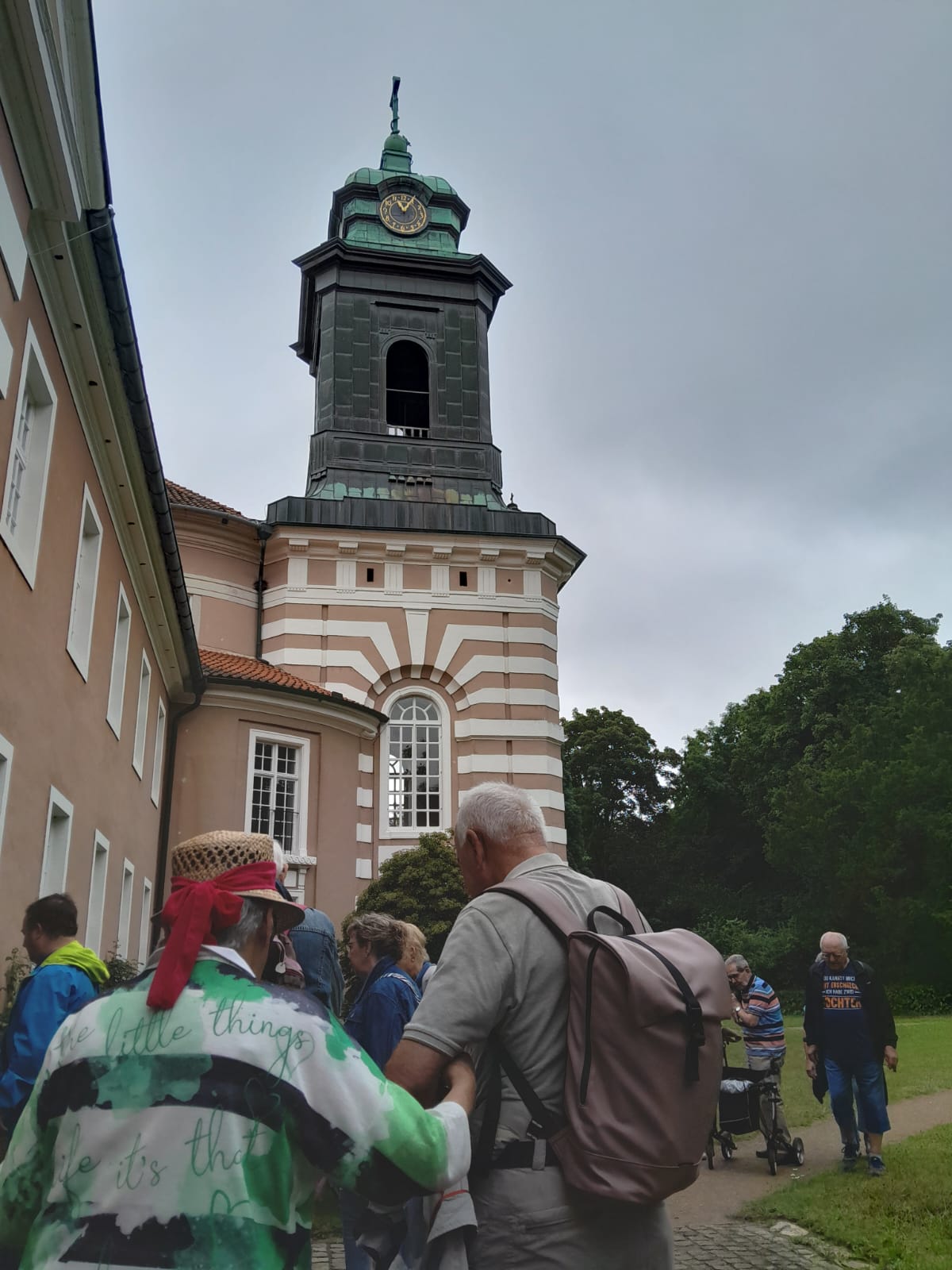 Einige der Reisenden stehen vor einem Glockenturm. Die Uhr zeigt 11:05 Uhr. Auf der rechten Seite schlie�t sich ein Park an das Geb�ude an.